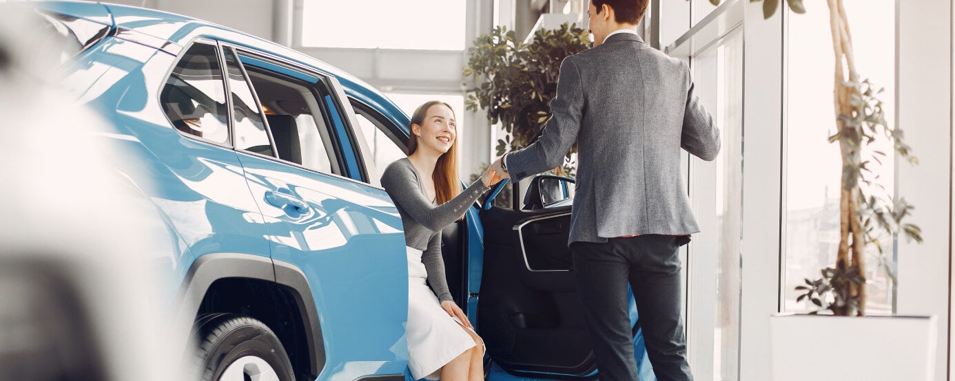 Dealership salesperson show the interior of a vehicle to a customer seated in a blue SUV inside the showroom.