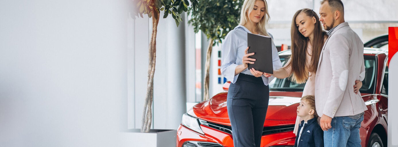Dealership salesperson reviews vehicle options with a family beside a red SUV in the showroom.