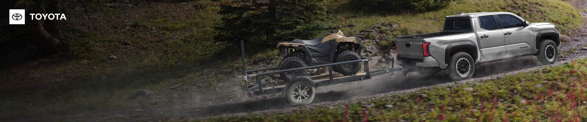 Rear view of a Toyota Tacoma TRD Off-Road in Celestial Silver towing an ATV trailer uphill on a mountain.