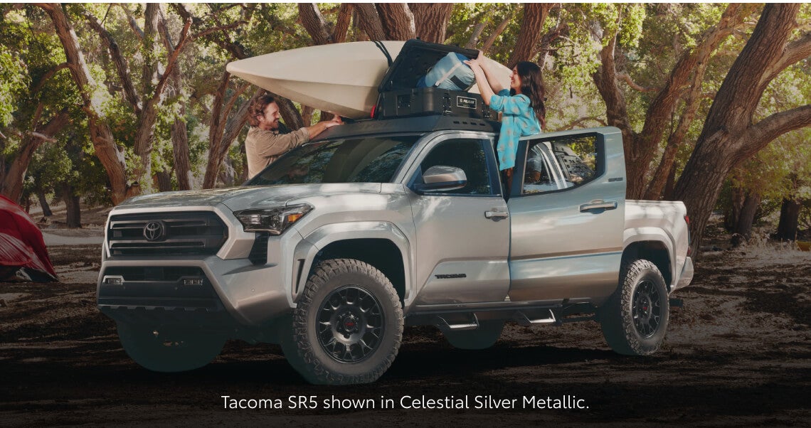 Front three-quarter view of a Toyota Tacoma SR5 in Celestial Silver parked in a wooded camp setting.
