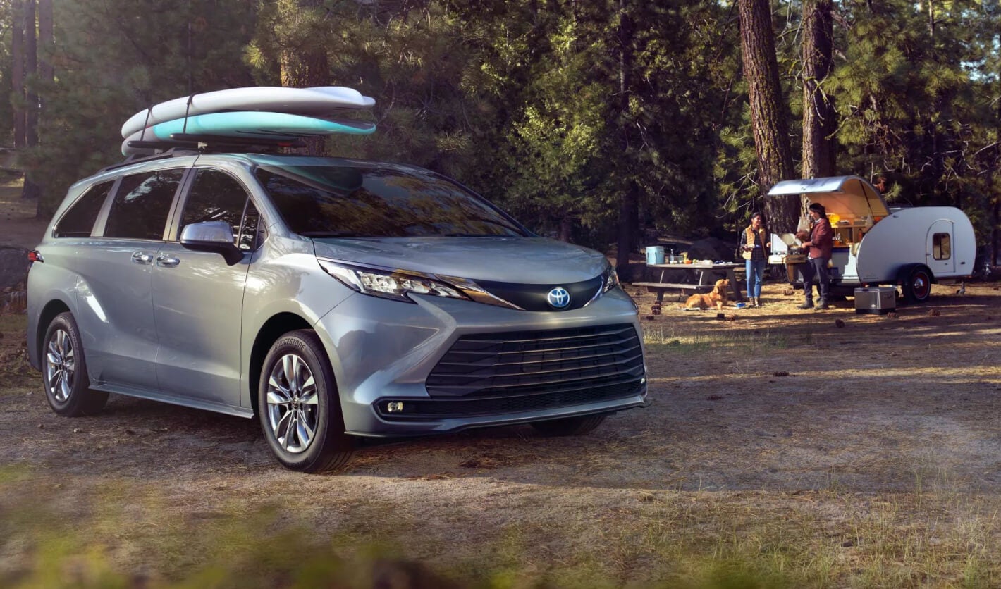 A gray Toyota Sienna parked at a campsite with trailer and kayaks strapped to roof rack.